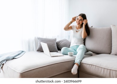 Woman enjoying a cup of coffee while relaxing with her laptop in living room. Female sitting on sofa with headphones watching movie on laptop. Smiling woman listening music in headphone from laptop