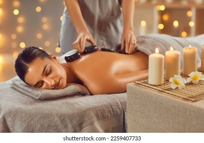 Woman enjoying exotic hot stones spa massage. Relaxed young woman lying on a spa bed while the masseuse is putting hot stones on her back. Spa treatment concept