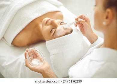 Woman enjoying facial treatment at beauty parlor or spa salon. Professional beautician holding small bowl with white clay mask and using brush to apply it on face of young woman client - Powered by Shutterstock