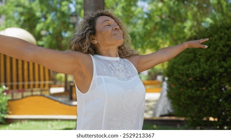 Woman enjoying freedom in a sunny park, with arms outstretched, wearing a white top, surrounded by greenery and urban elements, capturing a moment of relaxation and joy outdoors. - Powered by Shutterstock