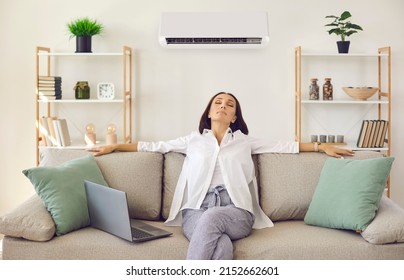 Woman Enjoying Modern AC System In Her Home. Happy Relaxed Middle Aged Lady Breathing In Fresh Air While Sitting On Comfortable Sofa With Laptop Computer In Living Room With Air Conditioner On Wall