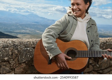 A woman enjoying playing guitar outdoors against a stunning mountain backdrop. She's smiling and dressed casually, expressing joy and relaxation in a serene natural setting. - Powered by Shutterstock