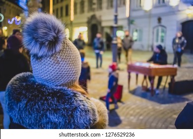 Woman Enjoying A Street Performance At Night While Young Kids Are Throwing Coins In The Bag Of A Street Performer On A Cold Busy Winter Street.