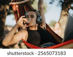 Woman enjoying summer in nature, relaxing on a hammock with a glass of wine and listening to music. Outdoor leisure and relaxation concept.