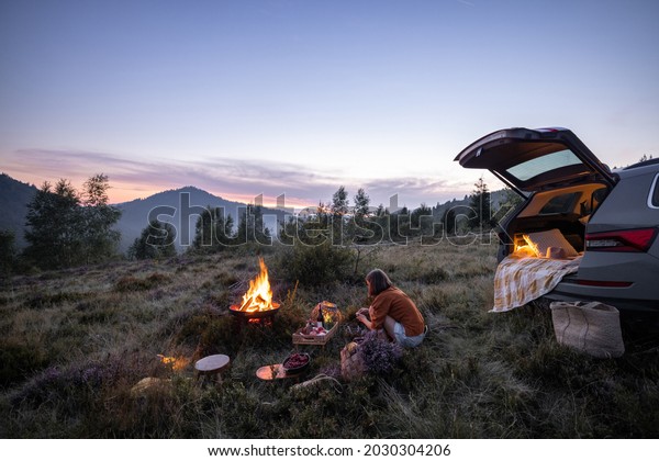 Woman enjoys beautiful view on the\
mountains, having a picnic with fireplace near the vehicle trunk at\
dusk on the evening. Traveling by car in\
nature
