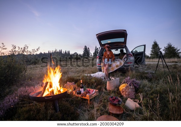 Woman enjoys beautiful view on
the mountains, having a picnic with fireplace and sitting in the
vehicle trunk at dusk on the evening. Traveling by car in
nature
