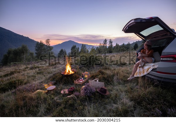 Woman enjoys beautiful view on\
the mountains, having a picnic with fireplace and sitting in the\
vehicle trunk at dusk on the evening. Traveling by car in\
nature