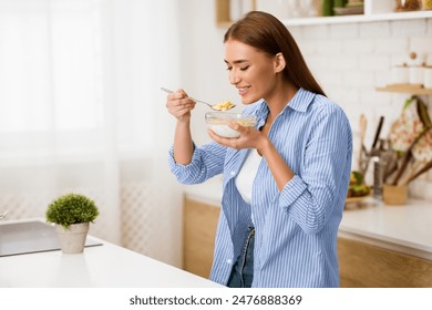 A woman enjoys a bowl of cereal in her kitchen, taking a bite with a spoon. - Powered by Shutterstock