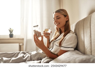 A woman enjoys her morning in bed, sipping water and taking her medication, embodying selfcare and wellness. This scene illustrates positive morning routines for health and happiness - Powered by Shutterstock