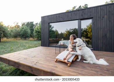 Die Frau genießt die Natur, während sie auf der Holzterrasse neben dem modernen Haus mit Panoramafenstern in der Nähe des Pinienwaldes sitzt und ihr Haustier umarmt. Konzept der Einsamkeit und Erholung in der Natur