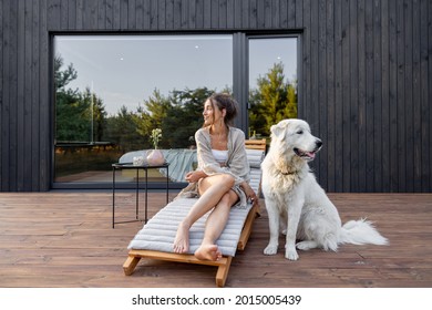 Woman enjoys the nature while sits on sunbed on wooden terrace at the modern hotel resort with panoramic windows near pine forest whith big white dog. Concept of solitude and recreation on nature