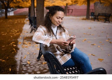 A woman enjoys a peaceful moment in a park, checking her smartphone amidst golden autumn leaves. She's relaxed and content, embodying a modern lifestyle in a natural setting. - Powered by Shutterstock