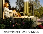 A woman enjoys a peaceful moment sitting by a large window, surrounded by blooming flowers in a sunlit garden. The scene captures the essence of escaping to a serene house in nature