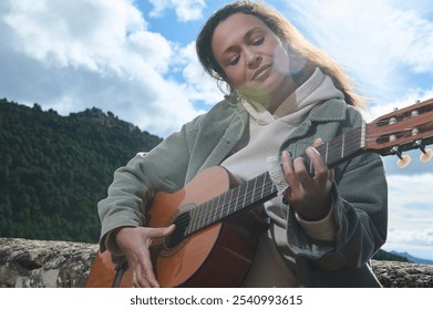 A woman enjoys playing a guitar with a scenic mountain view, embracing the peaceful outdoor atmosphere. Sunlight and nature create a serene, musical experience. - Powered by Shutterstock