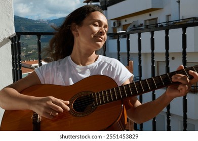 A woman enjoys playing her acoustic guitar on a balcony with clear blue skies and mountains in the distance. Her peaceful expression suggests relaxation and joy in music and nature. - Powered by Shutterstock