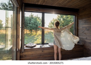 Woman enjoys sunrise in a country house or hotel staying with open hands near panoramic windows with pine forest view. Good morning and recreation on nature concept. Back view - Shutterstock ID 2015005271