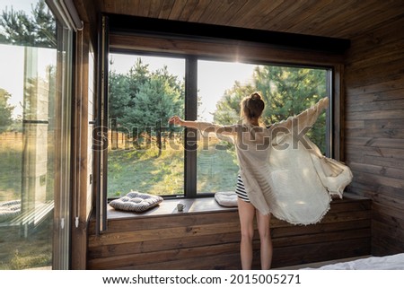 Woman enjoys sunrise in a country house or hotel staying with open hands near panoramic windows with pine forest view. Good morning and recreation on nature concept. Back view