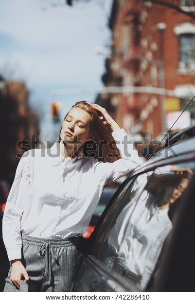 Woman\
enjoys the sunshine leaning to a car on the\
street