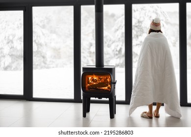 Woman enjoys winter time at home with burning fireplace and panoramic window, looking outside on snowy landscape. Concept of winter mood and comfort at home. Idea of winter vacation in the mountains