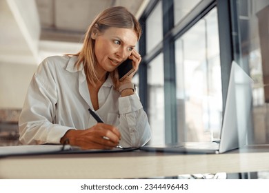 Woman entrepreneur making notes and talking by phone with client while sitting in coworking  - Powered by Shutterstock