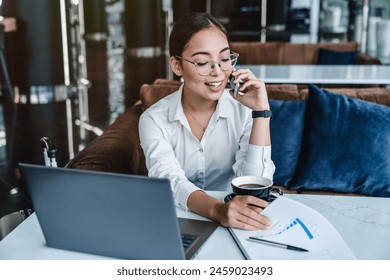 Woman entrepreneur managing her business from office lobby working on laptop computer and discussing business using smartphone drink cup of coffee. Businesswoman wearing suit making call on cellphone - Powered by Shutterstock