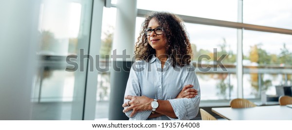 Woman entrepreneur standing beside a window in
office. Mature woman standing in office with arms crossed looking
outside window.