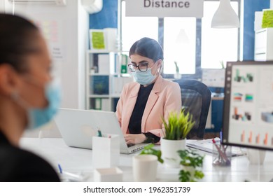 Woman Entrepreneur Working On Laptop In New Normal Office With Coworkers Keeping Social Distnacing Behind Plastic Shiled, Wearing Face Mask As Safety Prevention During Cornavirus Flue Outbreak.