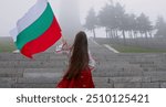 Woman in ethnic folklore costume with Bulgarian flag climbing Shipka monument, celebrating independence of the country