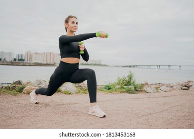 A Woman Of European Appearance, Playing Sports On The Street, Doing Exercises Squat Lunges Forward, Holding Small Dumbbells. Looks Ahead Thinks About Proper Nutrition