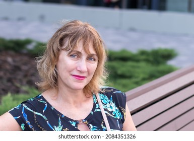 Woman, A European, Middle-aged, 50-55 Years Old, A Pretty Blonde, Sitting On A Park Bench With A Smile