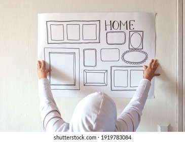 Woman Evaluating And Sizing Up White Poster With Drawn Set Of Different Empty Vertical And Horizontal Picture Frames On White Wall, Planning Family Photo Gallery Project To Capture Moment, Lifestyle