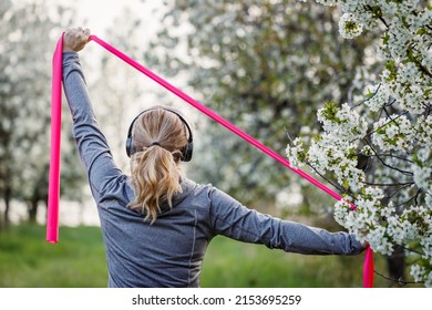 Woman exercising with elastic resistance band in spring nature. Sportswoman with headphones listening music during fitness and warm up exercise