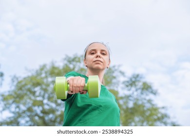 Woman exercising with a green dumbbell outdoors during a sunny day in a park setting. - Powered by Shutterstock