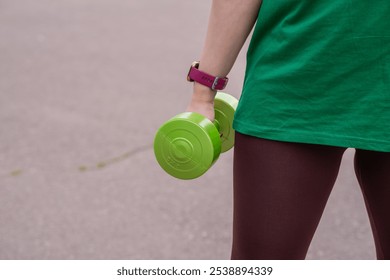 Woman exercising with a green dumbbell outdoors during a sunny day in a park setting. - Powered by Shutterstock