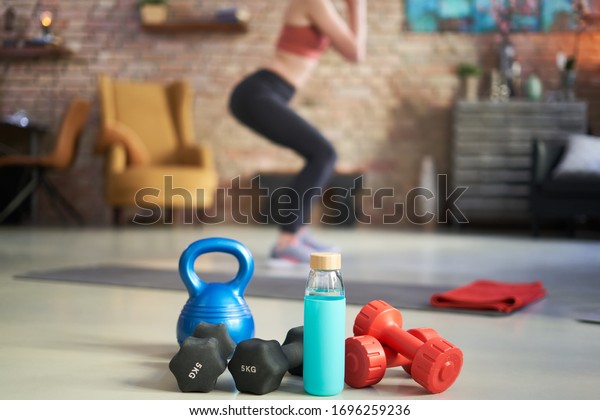 Woman exercising at home in living room, focus on\
fitness equipments, barbell and kettlebell. Woman doing squats in\
the background. Concepts about home workout, fitness, sport and\
health.