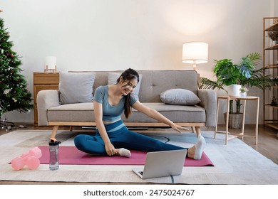 Woman Exercising at Home in Modern Living Room, Stretching on Yoga Mat with Laptop, Water Bottle, and Dumbbells, Cozy and Bright Interior with Sofa, Plants, and Christmas Tree - Powered by Shutterstock