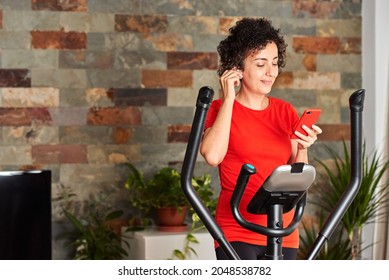 Woman Exercising At Home On Cross Training Elliptical Using Mobile Phone