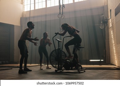 Woman Exercising On Air Bike At Gym With Friends Motivating. Female Using Air Bike For Workout At Cross Training Gym With Friends.