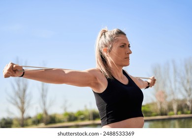 A woman is exercising outdoors with a resistance band. She is wearing a black tank top and has her arms raised. The scene is bright and sunny, and the woman is enjoying her workout - Powered by Shutterstock