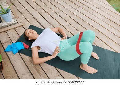 Woman exercising with a resistance band on a yoga mat outdoors, promoting fitness and relaxation on a wooden deck. - Powered by Shutterstock