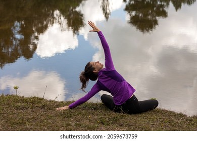 Woman Exercising Yoga Outdoors In Front Of A Pond With Cloud Reflections In The Water.

