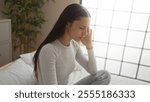 Woman experiencing headache sitting on bed in a bedroom with plants and natural light