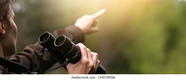 Woman explorer with binoculars showing with finger for animal in the wild forest. Blur dark background. The concept of hunting, travel and outdoor recreation.  - Powered by Shutterstock