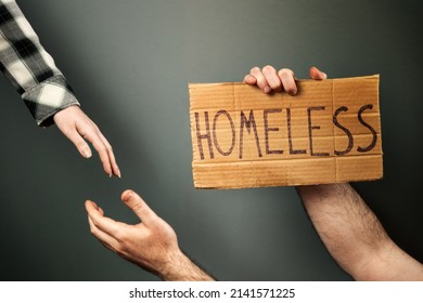 A woman extends a helping hand to a man holding a cardboard box with the text homeless. Dark background of a greenish hue. The concept of a social problem with people without a home and vagabonds.