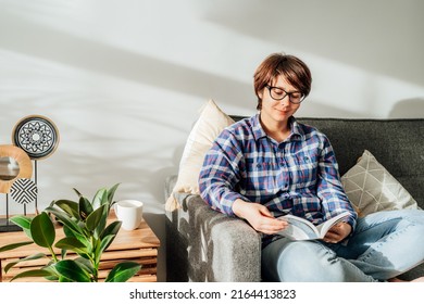Woman in eye glasses sit on the sofa and relax while reading a book at home in cozy modern interior living room with green plants. Offline activity lifestyle. Reading hobby. Leisure and entertainment