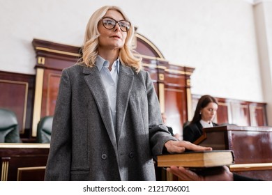 woman in eyeglasses giving swear on bible in court near prosecutor on blurred background