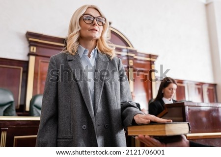woman in eyeglasses giving swear on bible in court near prosecutor on blurred background