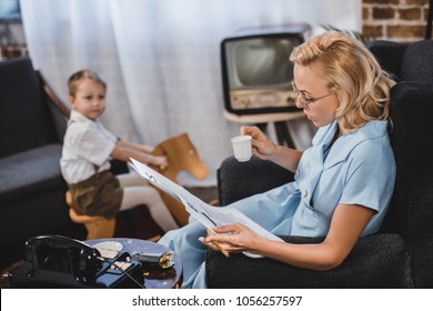 woman in eyeglasses holding cup of coffee and reading newspaper while little son sitting on rocking horse behind, 50s style family
