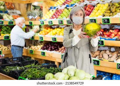 Woman In Face Mask Choosing Melon While Standing In Greengrocer. Old Man Shopping In Background.