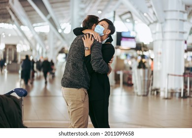 Woman In Face Mask Giving Welcome Hug To Man At Airport Arrival Gate. Woman Wearing Face Mask Welcoming And Embracing Her Boyfriend At Airport